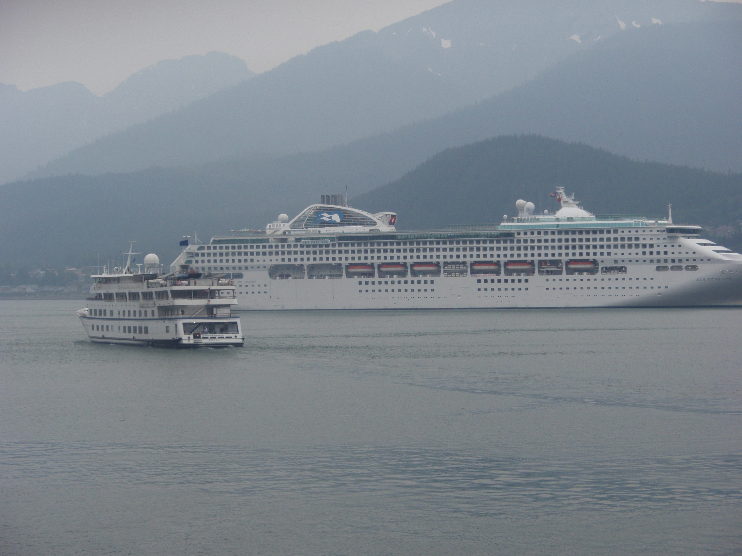 Phtos shows two cruise ships in a foggy bay. One very large, the other much smaller. Rows of hills inthe background