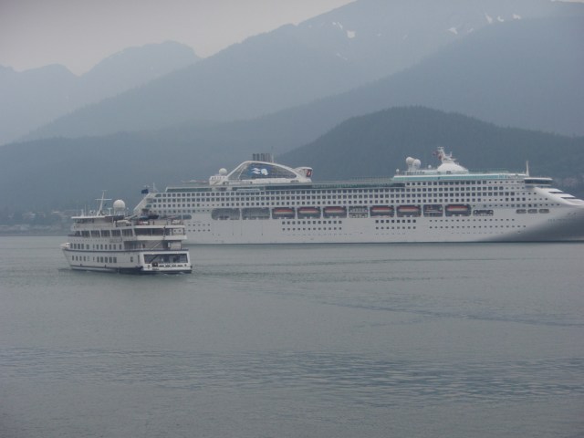 Phtos shows two cruise ships in a foggy bay. One very large, the other much smaller. Rows of hills inthe background