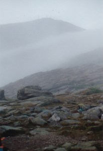 Photo shows the rocky foreground with the summit of Mount Washington, NH, in the background and in fog
