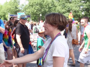 Photo shows Maura Healey, a white woman with short hair and a white T-shirt and Pride beads, holding out her hand to someone, in 2015 at Boston's Pride march.