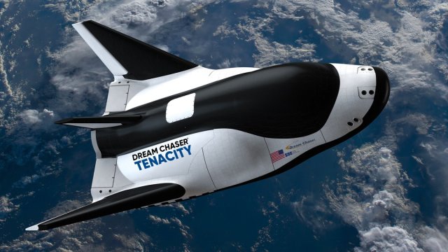 Artist representation of a small cargo space plane with Earth and clouds below. Black and white with wings. Name on the side: Dream Chaser Tenacity. Small American flag on the side.