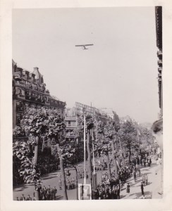Black and white photo of a military parade in Paris, France, on VE Day, 1945. With an airplane flying overhead.