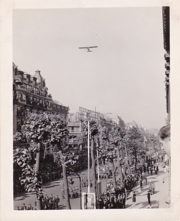 Black and white photo of a military parade in Paris, France, on VE Day, 1945. With an airplane flying overhead.