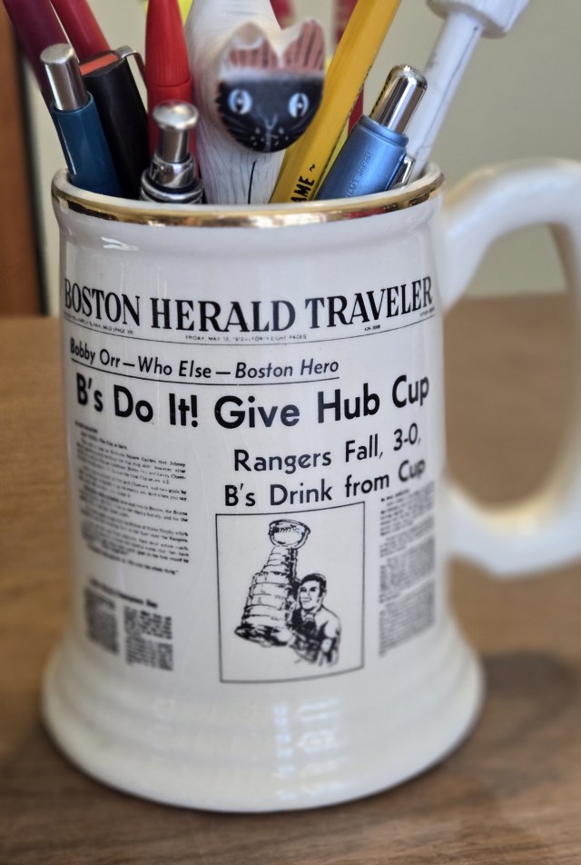 Photo of a ceramic mug with a replica of the Boston Herald Traveler front page: B's Do It! Give Hub Cup" from 1972. Rangers fall, 3-0. B's Drink from Cup. Image is Johnny Bucyk holding the Stanley Cup.